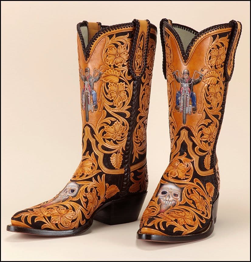 A pair of tan and black cowboy boots carved with scrolling foliage, a motorcycle rider on each shaft and a skull at the toe.