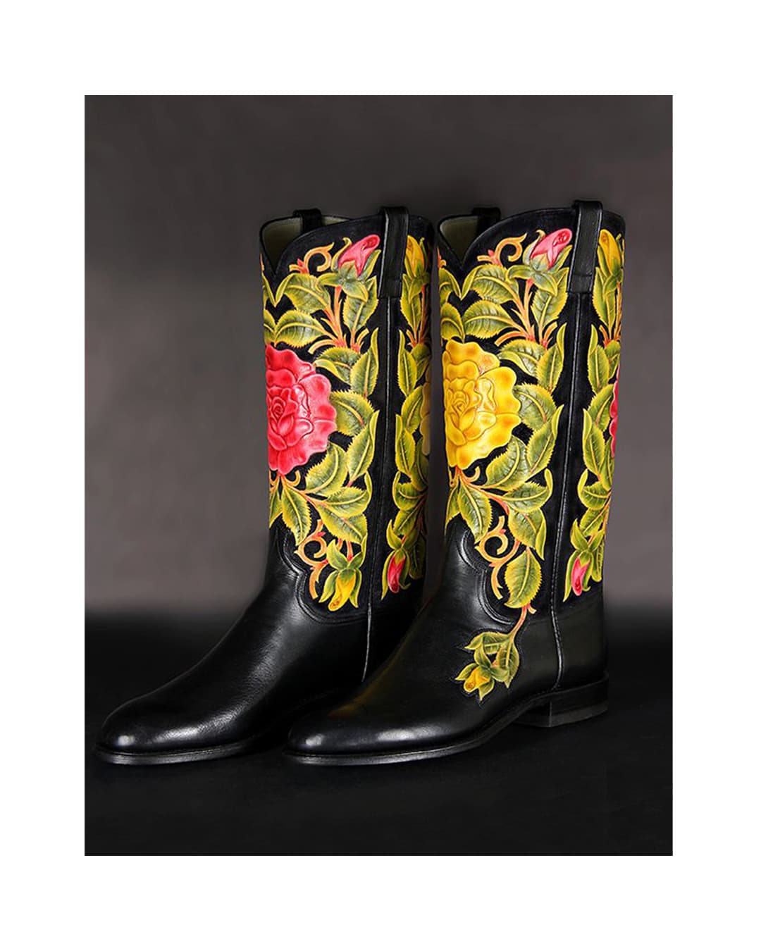 A pair of black cowboy boots with tops carved and coloured with pink and yellow roses among green leaves.