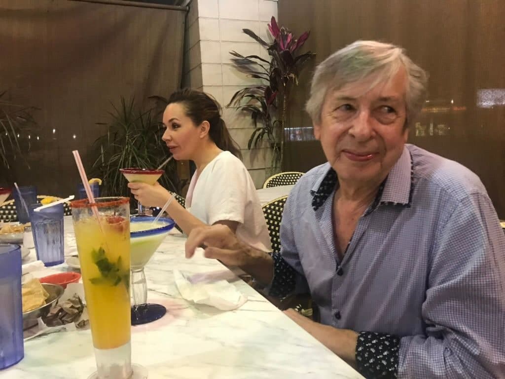 Peter at a marble table turning to the camera with a half-smile, cocktails and bowls of crisps in front of him, his daughter beside him raising a glass.
