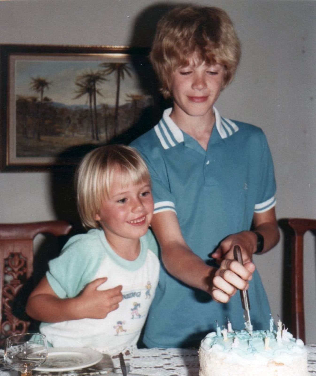 Two fair-haired children at a dining table cutting a pale blue birthday cake covered in candles, a framed painting of palm trees on the wall behind them.