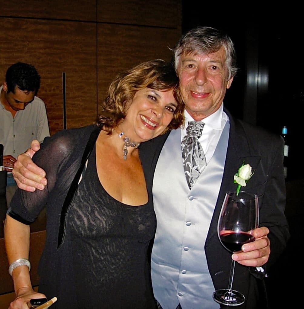 Peter in a dinner suit, waistcoat and cravat with a white rose in his lapel, a glass of red wine in one hand and his other arm around a smiling woman in black.