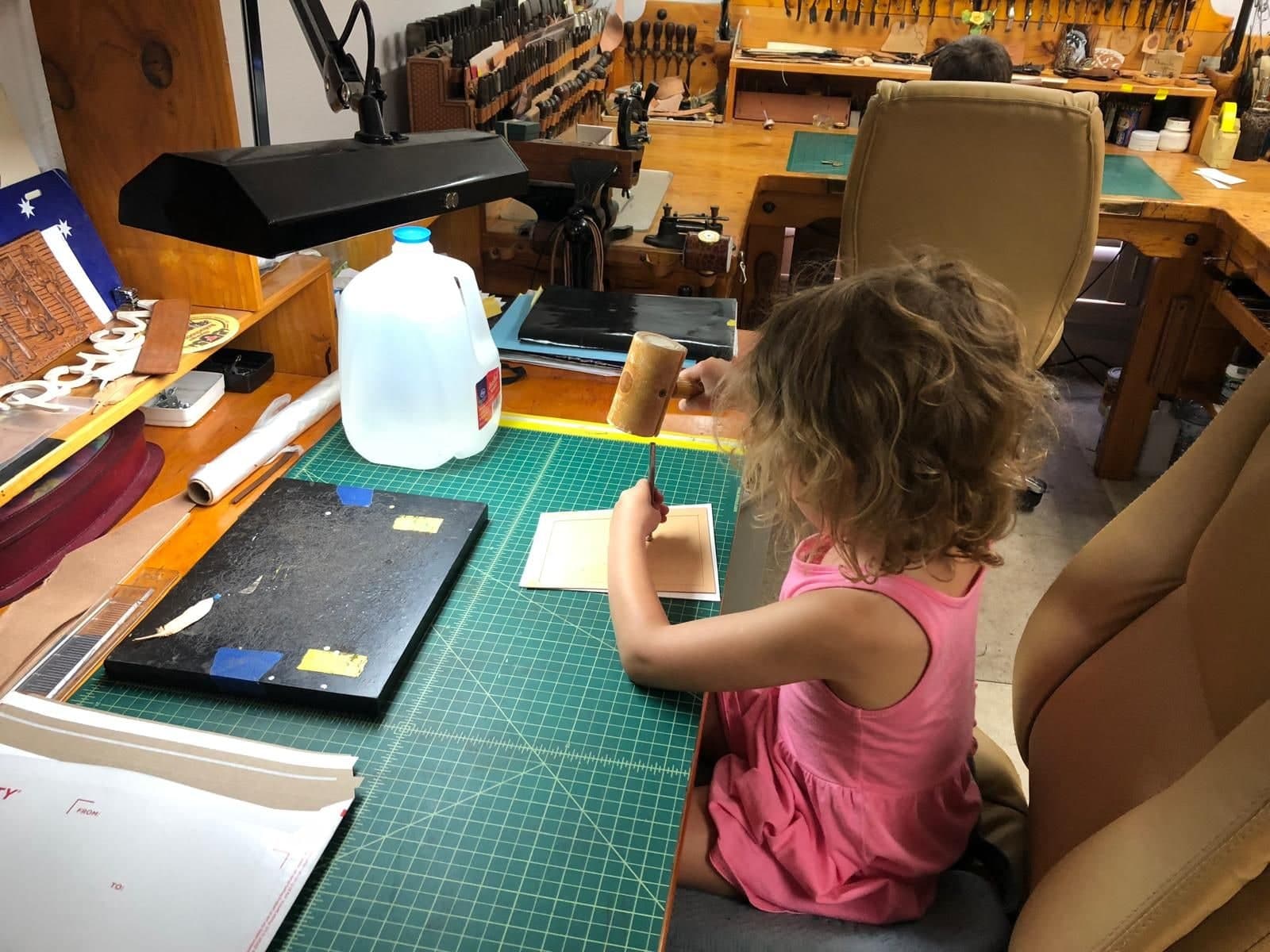 A small girl in a pink dress kneeling up at Peter’s workbench, mallet raised over a stamping tool on a piece of leather, the workshop stretching away behind her.