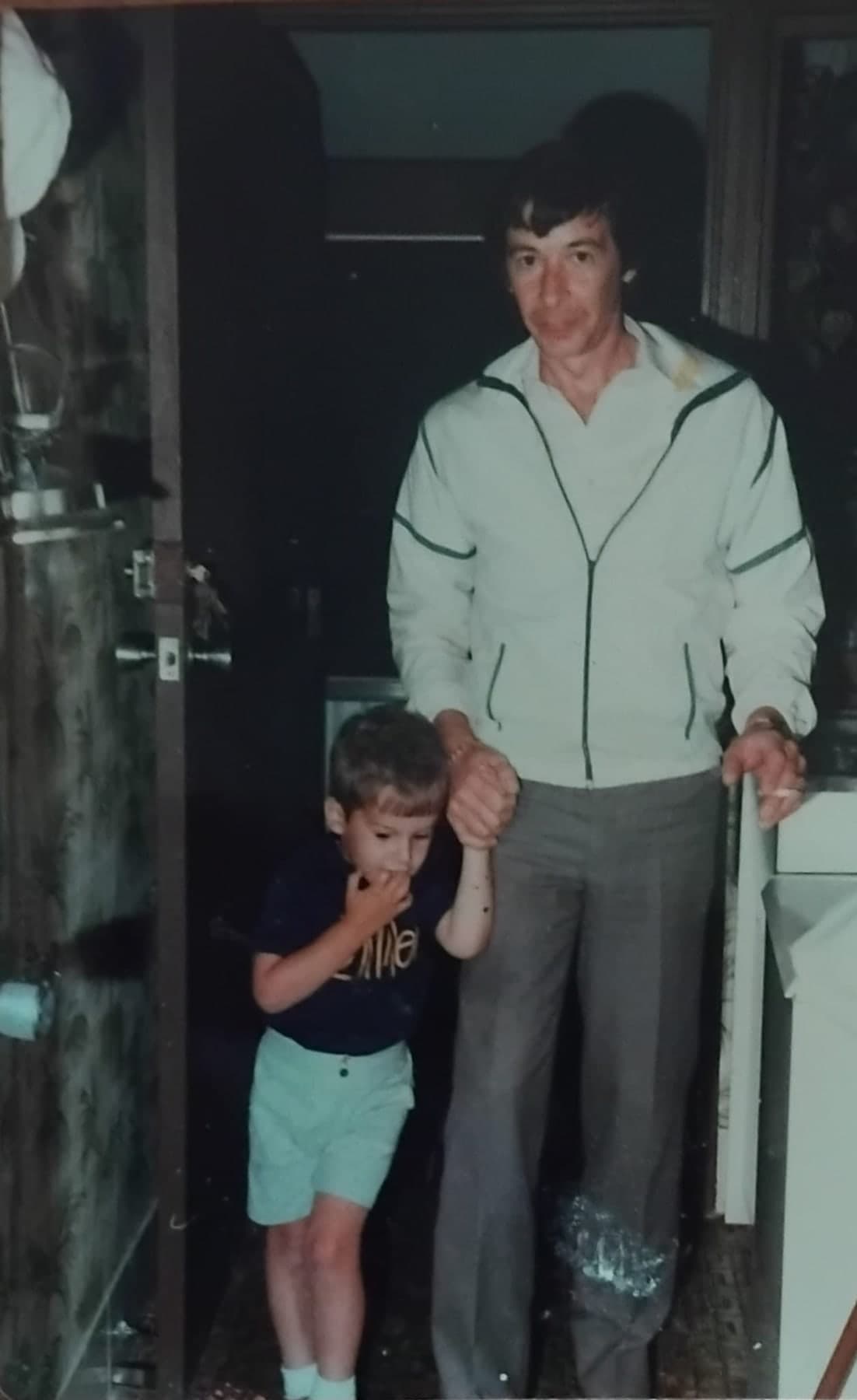 Peter in a pale zip jacket standing in a doorway, holding the hand of a small boy in a dark t-shirt and shorts.