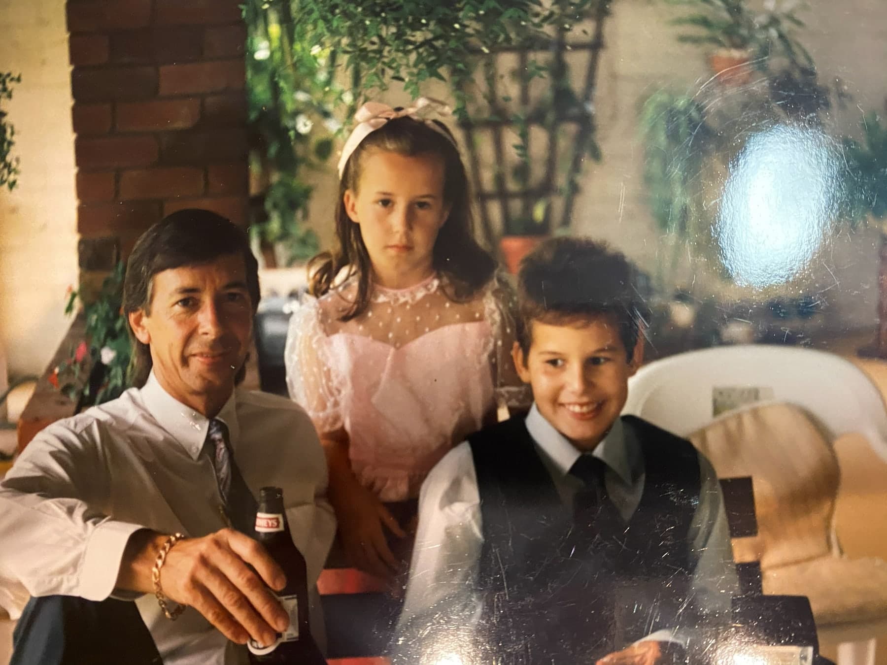 Peter in shirtsleeves and a tie, sitting with Emma in a pink party dress and Matthew in a waistcoat and tie, in front of a brick fireplace and trailing plants.