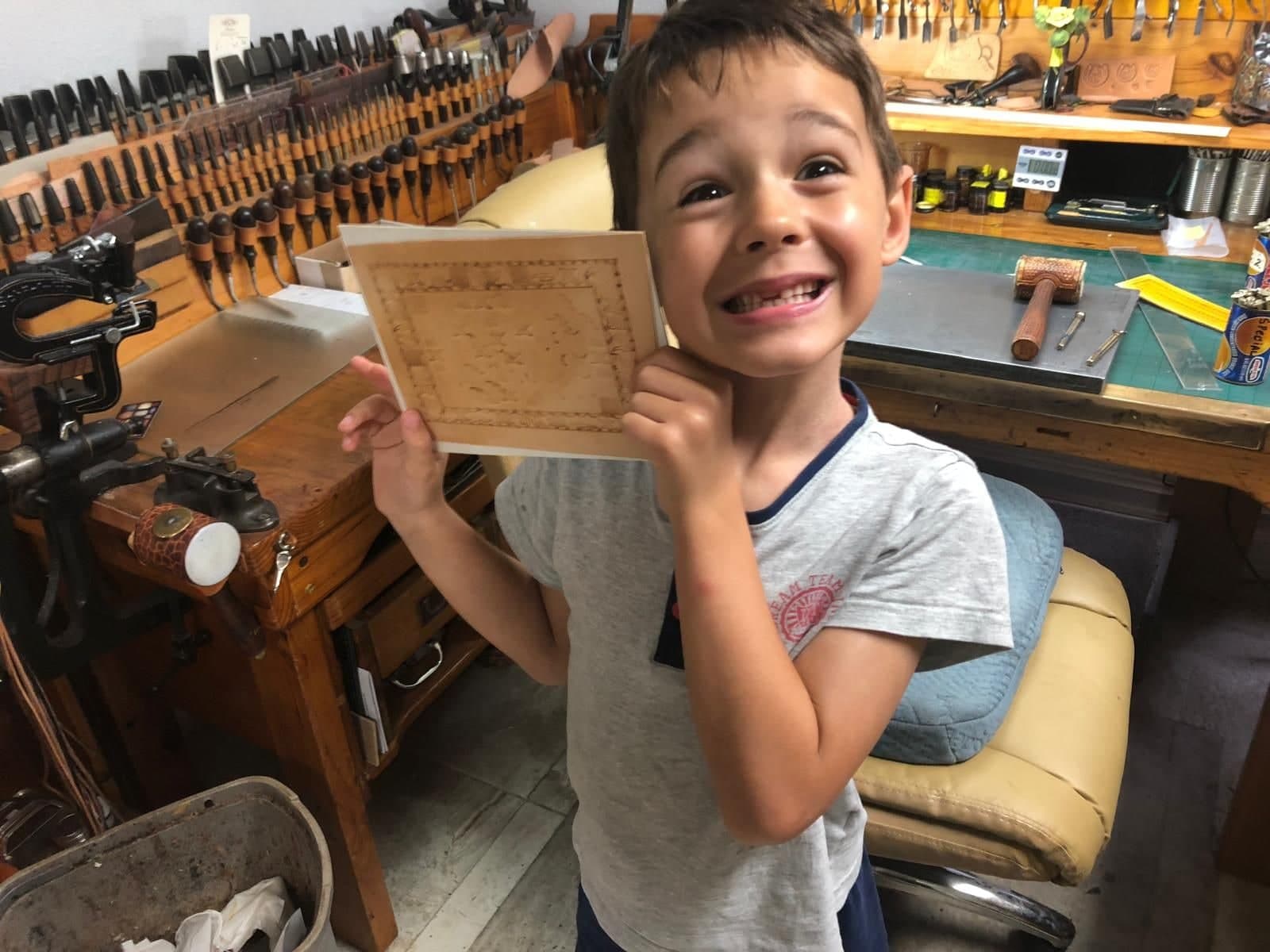 A boy in the workshop grinning widely, holding up a piece of tan leather he has tooled with a decorative border.