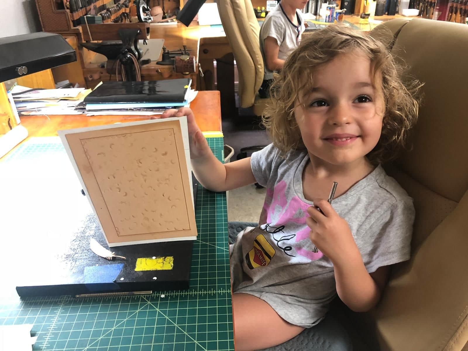 A small girl with curly fair hair sitting at the bench, holding up a piece of tan leather covered in the marks she has stamped into it, a tool still in her other hand.