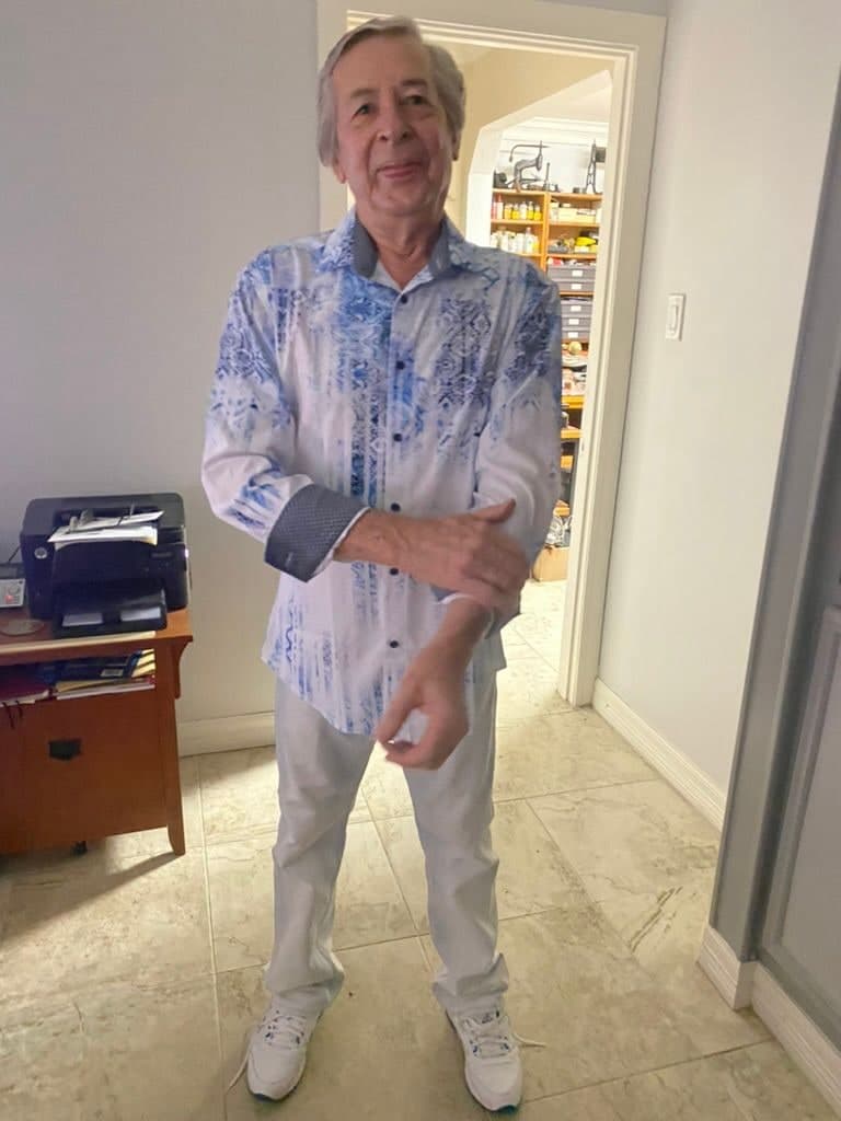 Peter standing in a hallway in a blue patterned shirt and pale trousers, mid-sentence, the door to his workshop open behind him.