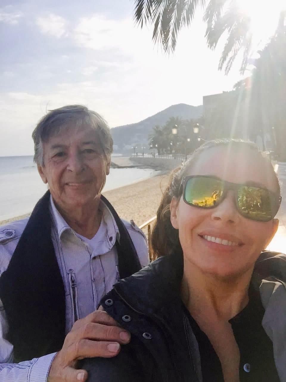 Peter with his hand on his daughter’s shoulder on the beach at Ibiza at golden hour, the bay and the headland behind them.