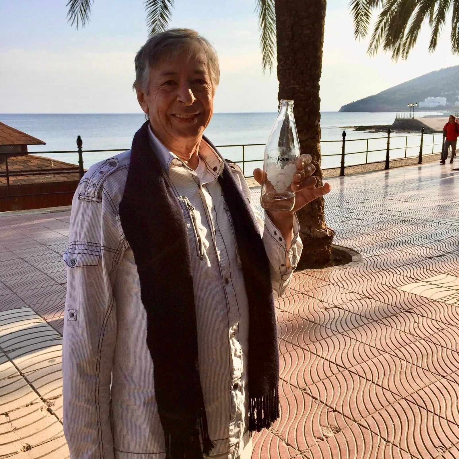 Peter on a seafront promenade in Ibiza in a pale jacket and dark scarf, holding up a glass bottle of water, palm trees and the sea behind him.