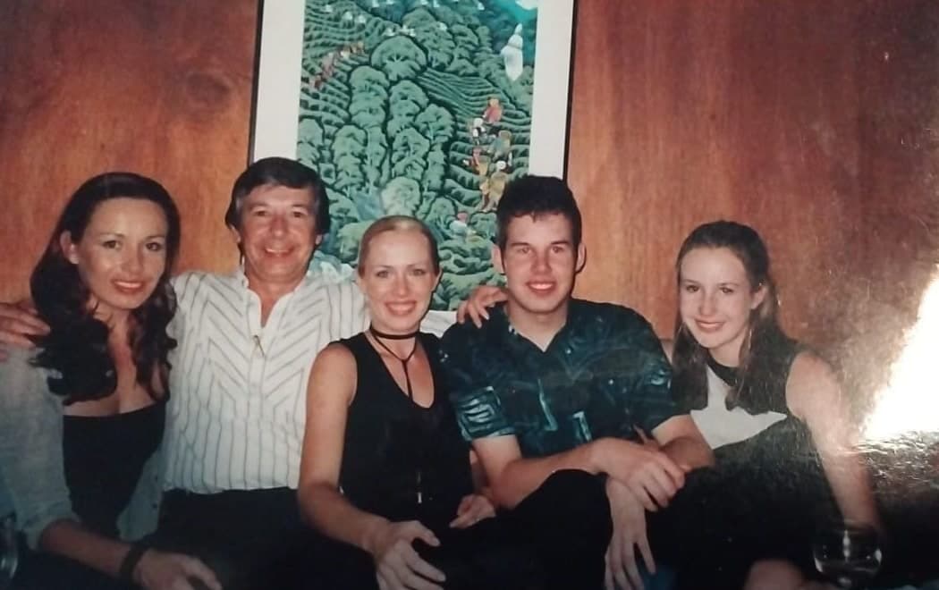 Peter in a striped shirt sitting on a sofa between his four grown children, all leaning into one another, a large framed painting on the wall behind.