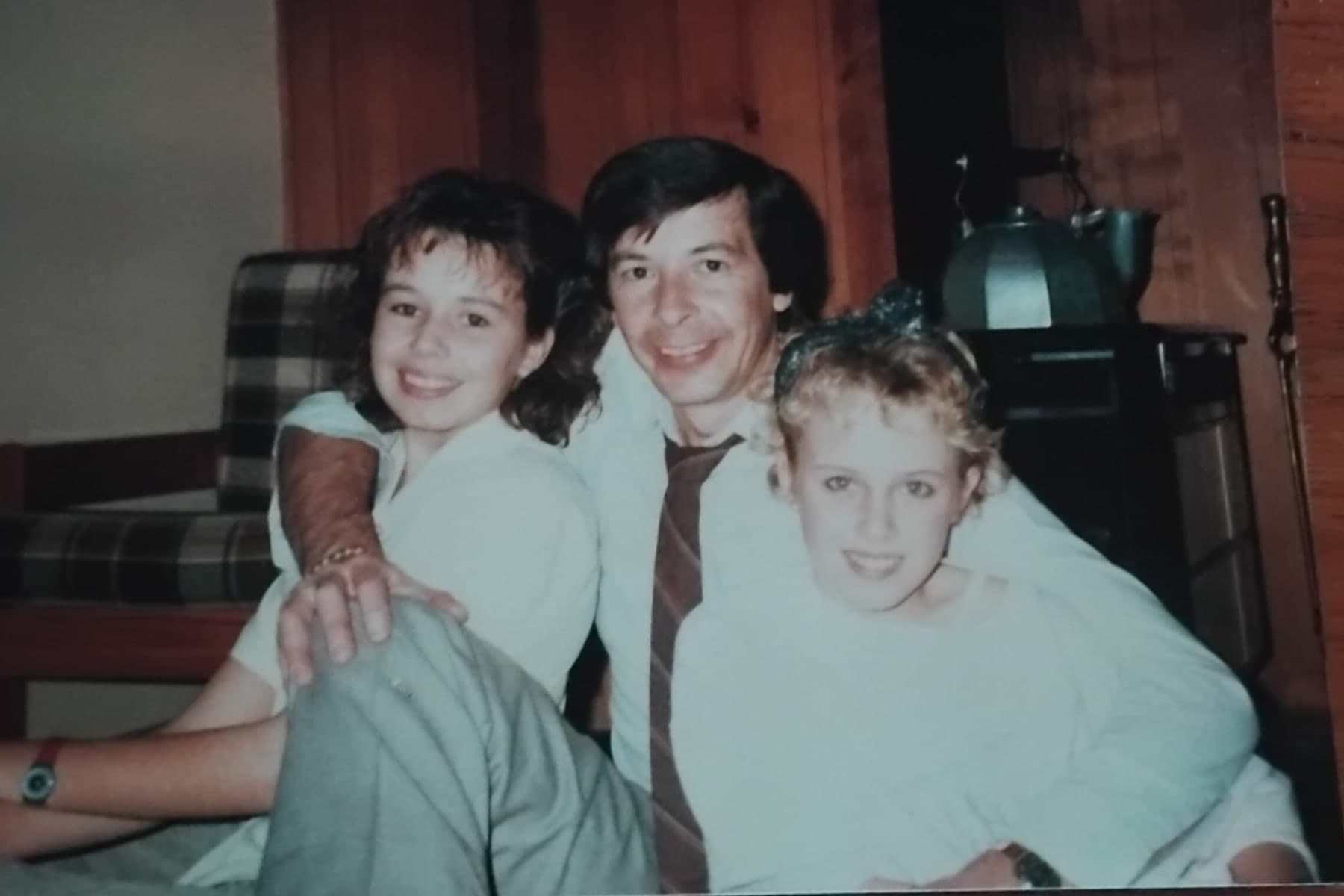 Peter in a shirt and striped tie sitting between Pietra and Sarah as teenagers, all three leaning in and smiling, a timber-lined room behind them.
