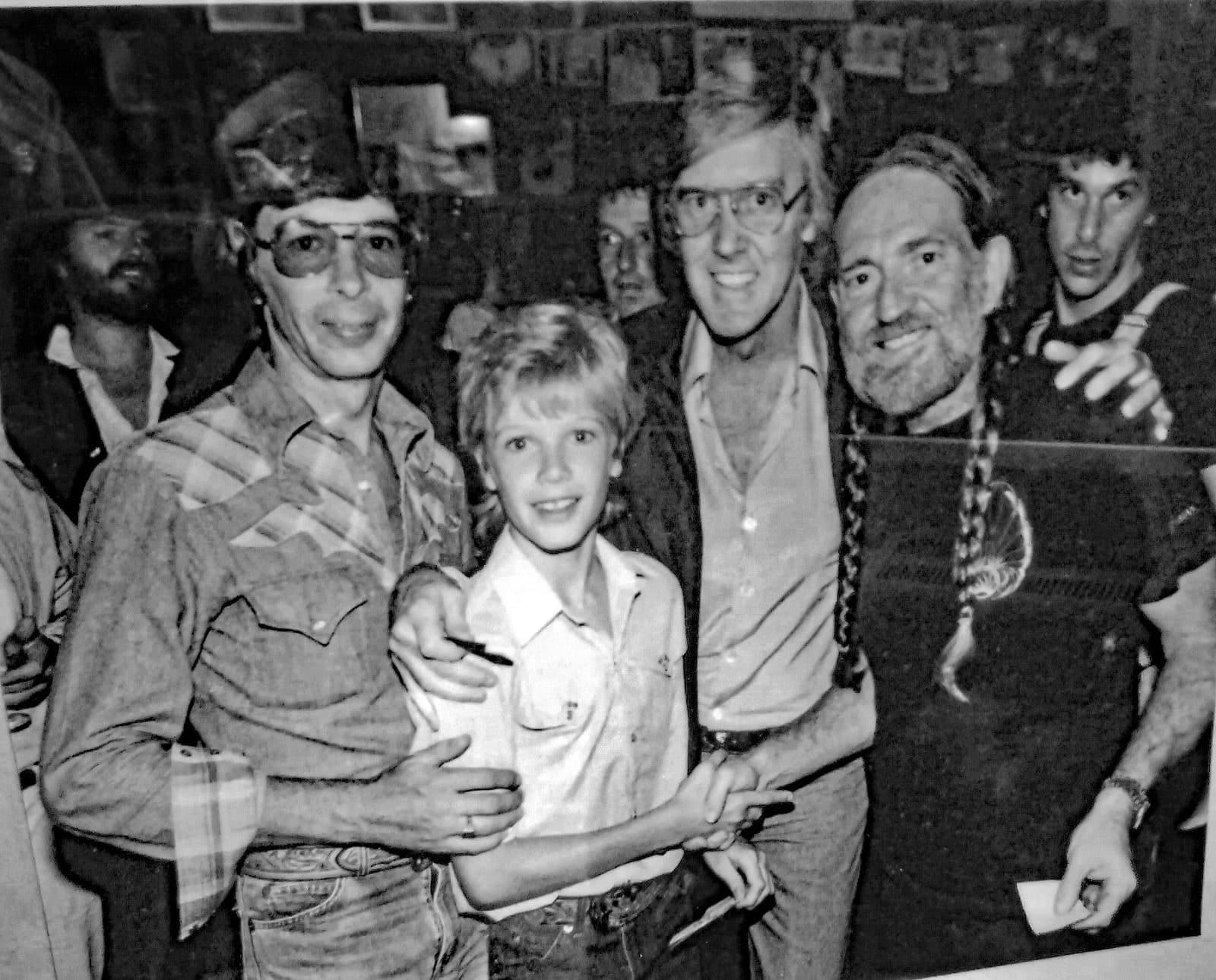 A black and white photograph of a crowd backstage. A framed leather panel is being held up at the right of the group, and a young person stands at the centre with an arm around them.