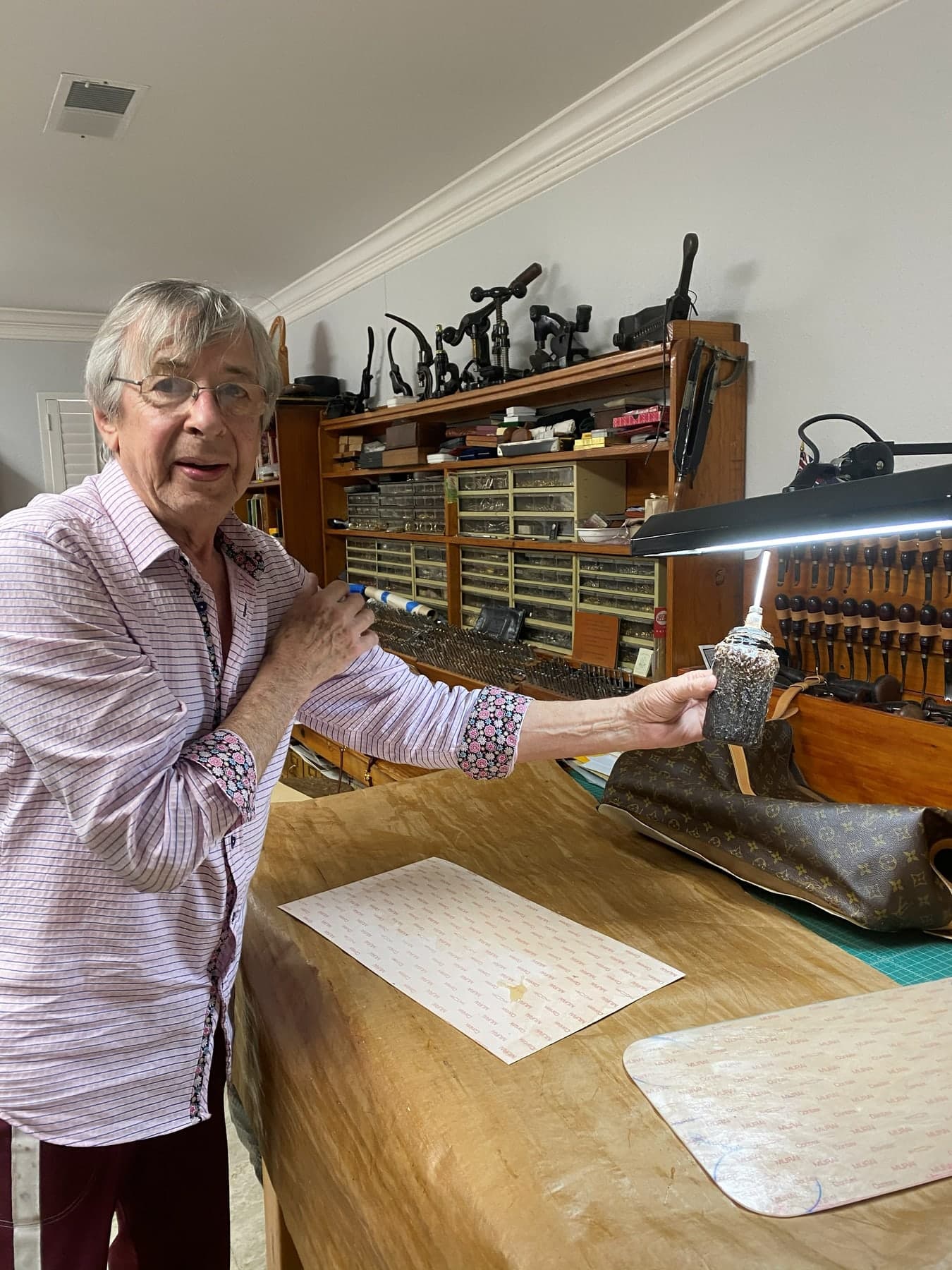 Peter in a pink striped shirt in his Houston studio, holding up a small carved piece, racks of stamping tools and drawers of fittings behind him.