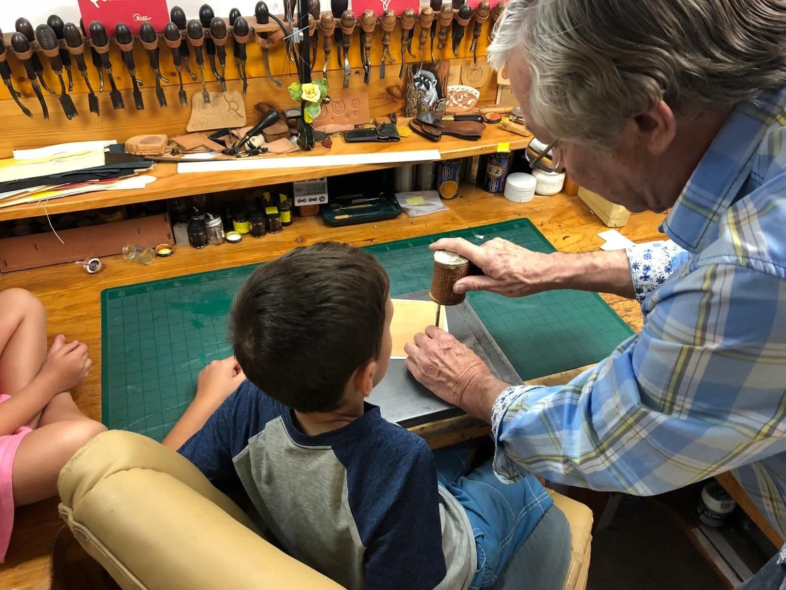 Seen from above: Peter in a blue checked shirt leaning over a small boy at the workbench, guiding a stamping tool and mallet onto a piece of leather, the whole bench and its racked tools spread out in front of them.