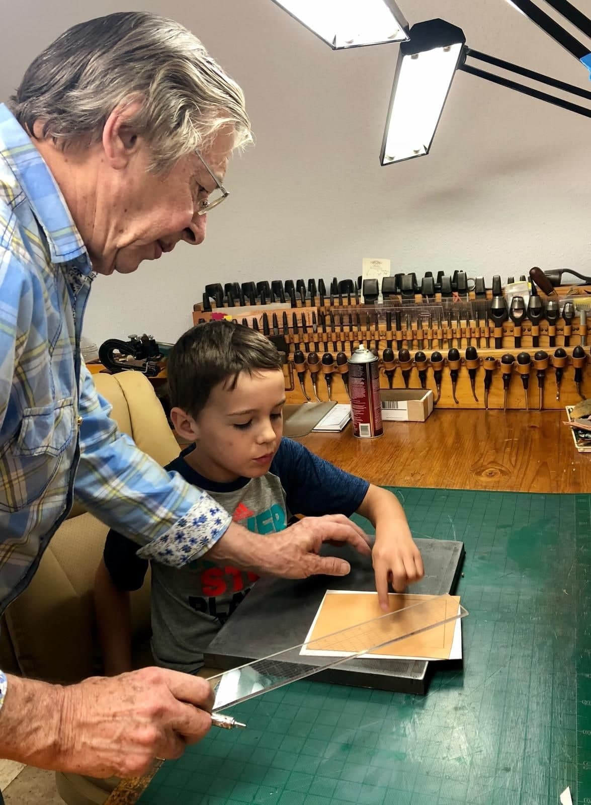 Peter bending over the bench beside a young boy, drawing a border line along a piece of tan leather with a steel rule, hundreds of stamping tools racked on the wall behind.