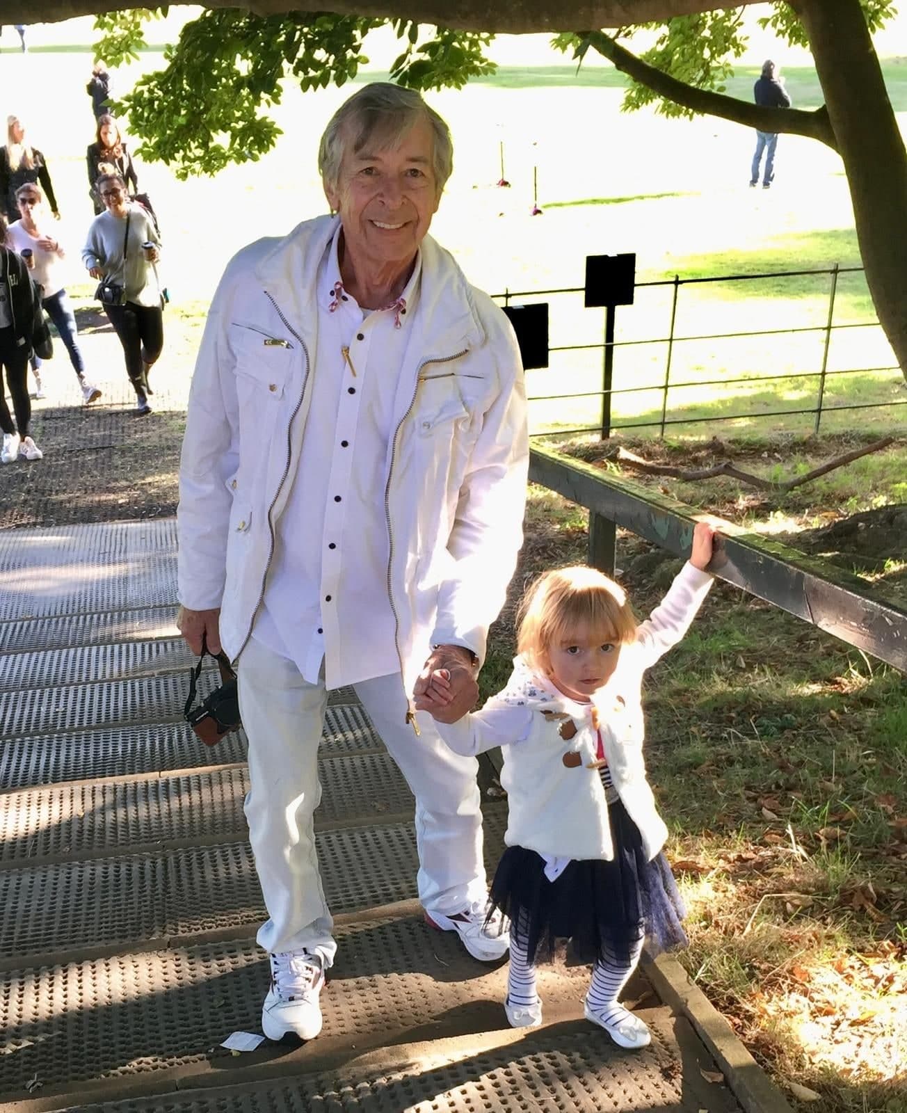 Peter dressed head to toe in white, camera in one hand, holding the hand of a small blonde child in a white cardigan and navy skirt on a walkway through a park.