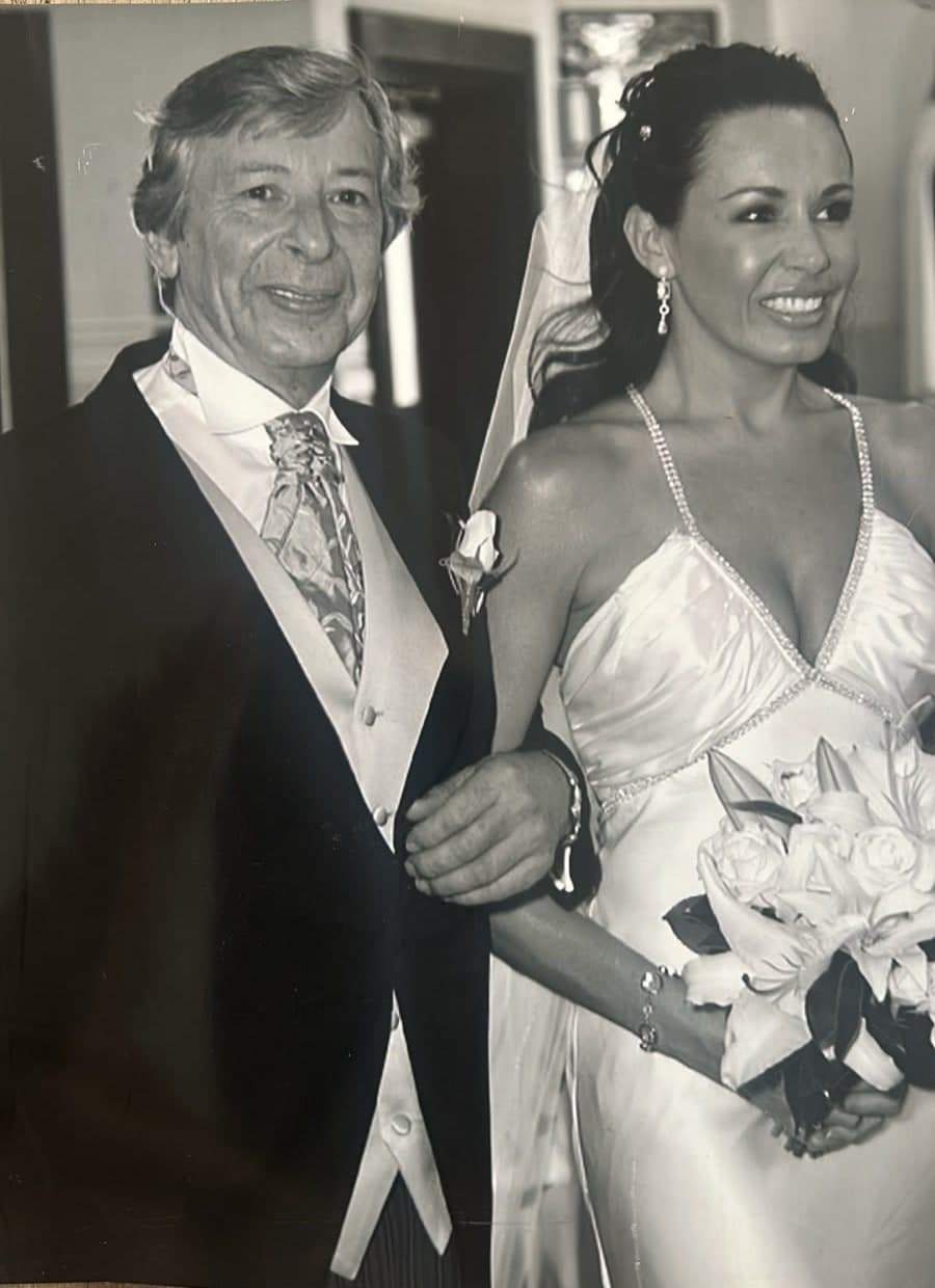 A black and white photograph of Peter in morning dress with a cravat and a white rose buttonhole, arm in arm with his daughter in her wedding dress and veil, holding a bouquet of white lilies and roses.