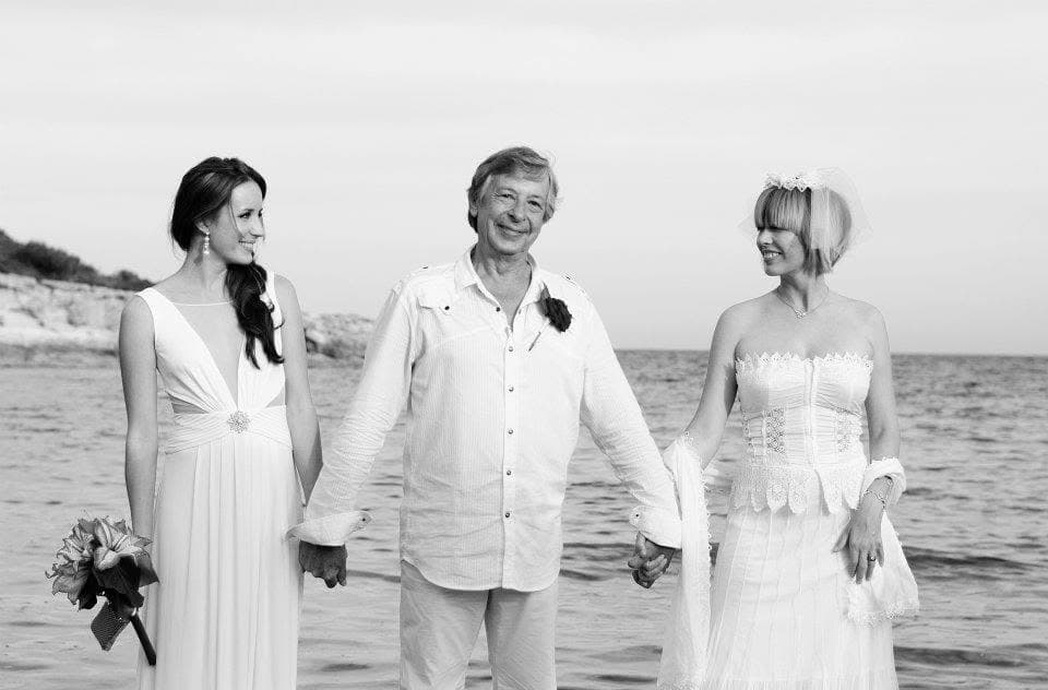 A black and white photograph of Peter on a beach in Ibiza, in a white shirt and trousers, smiling, holding hands with his daughters Sarah and Emma, both dressed in white, the sea behind them.