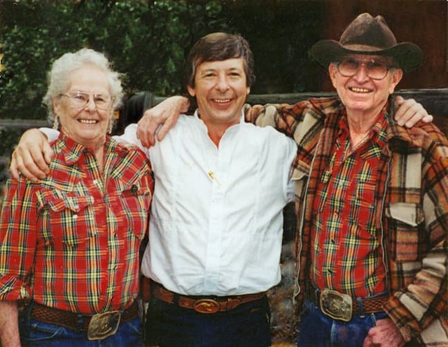 Peter in a white shirt with his arms around an older couple in checked shirts and tooled belt buckles, one of them in a wide-brimmed hat.