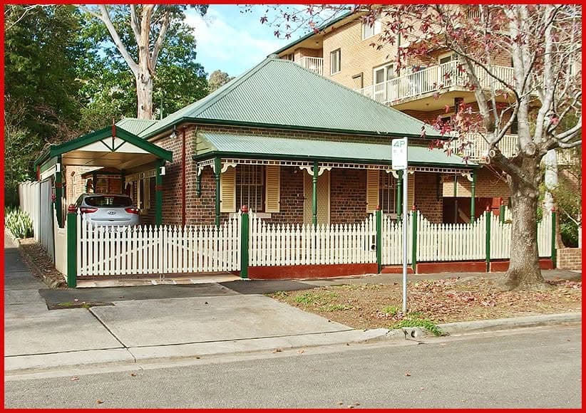 A brick cottage with a green corrugated roof, green posts and a white picket fence, on a Sydney street.