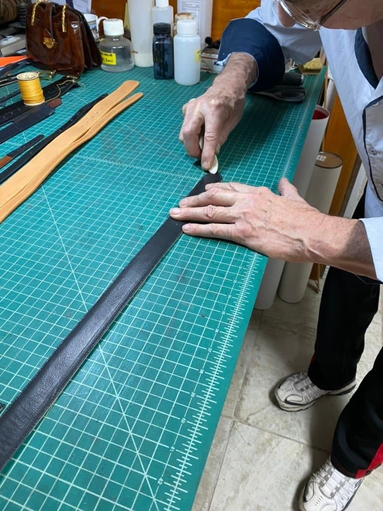 Peter’s hands at work on a long black leather belt laid out on a green cutting mat, finished belts and a leather handbag on the bench beyond.