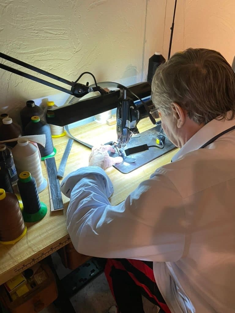 Peter bent over a stitching machine under a lamp, feeding leather through by hand, cones of thread in every colour racked along the bench beside him.