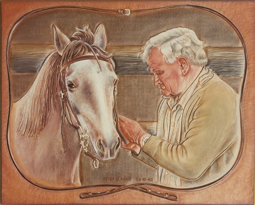 A carved and coloured leather picture of a white-haired man in a cardigan reaching up to hold the muzzle of a bridled grey horse, framed within a carved leather bridle and rein.