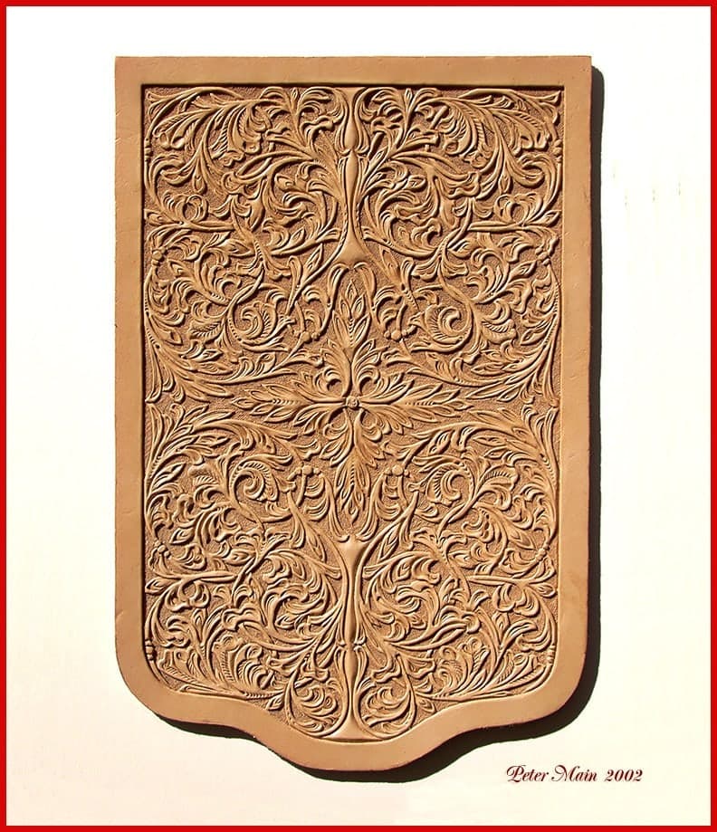 A large undyed leather panel with a shaped foot, carved edge to edge with dense symmetrical scrollwork and leaves, left in its natural tan so only the tooling gives it depth.