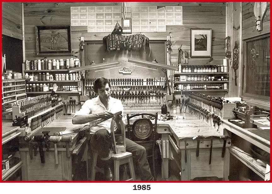 Peter at his workbench in 1985, tools racked in rows around him, working a long carved strap across his knee.