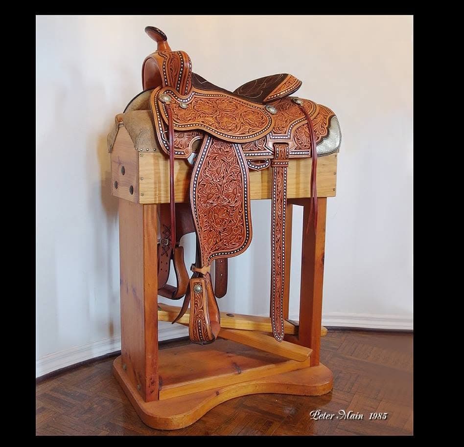 A hand-tooled Western saddle covered in floral carving with fine white edge stitching, on a timber stand.